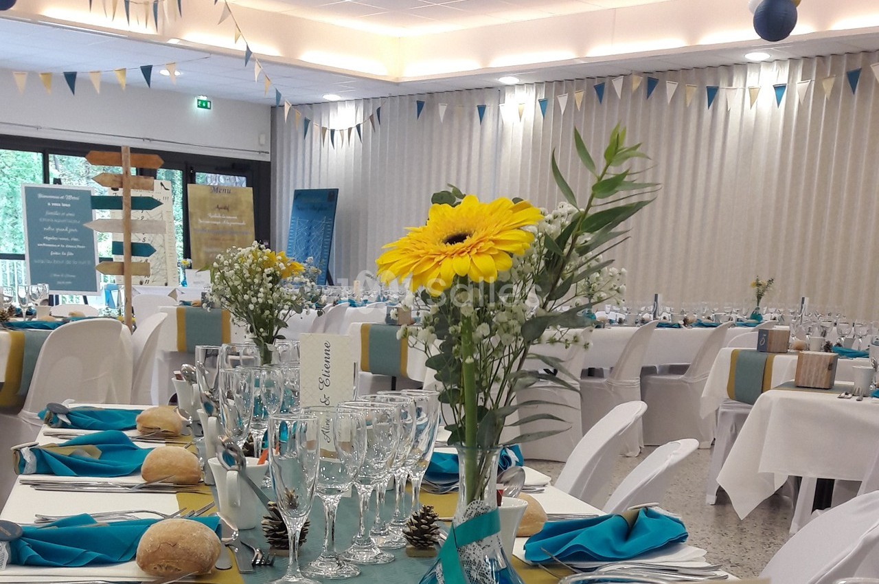 Salle de réception décorée pour un événement, avec tables dressées, nappes blanches, fleurs et serviettes bleues.
