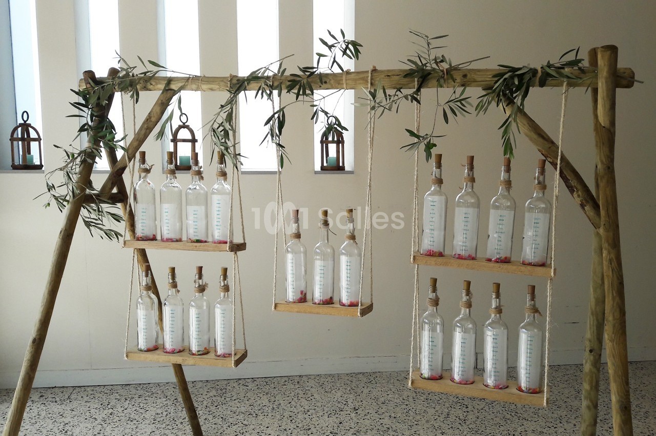 Bouteilles en verre transparent alignées sur des étagères suspendues à une structure en bois décorée de branches d'olivier.