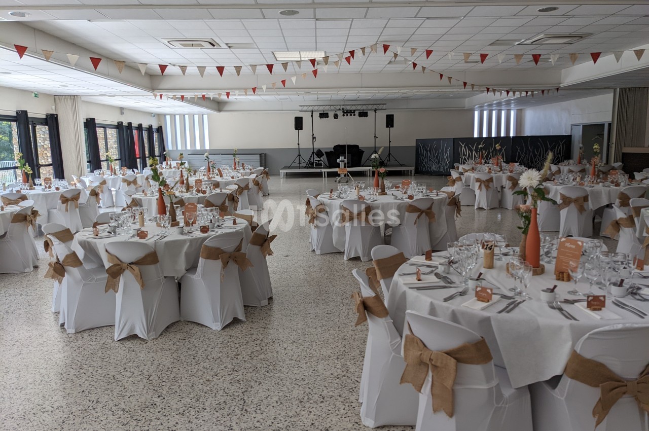 Salle de réception décorée avec des tables rondes dressées, nappes blanches et chaises ornées de nœuds en toile de jute.