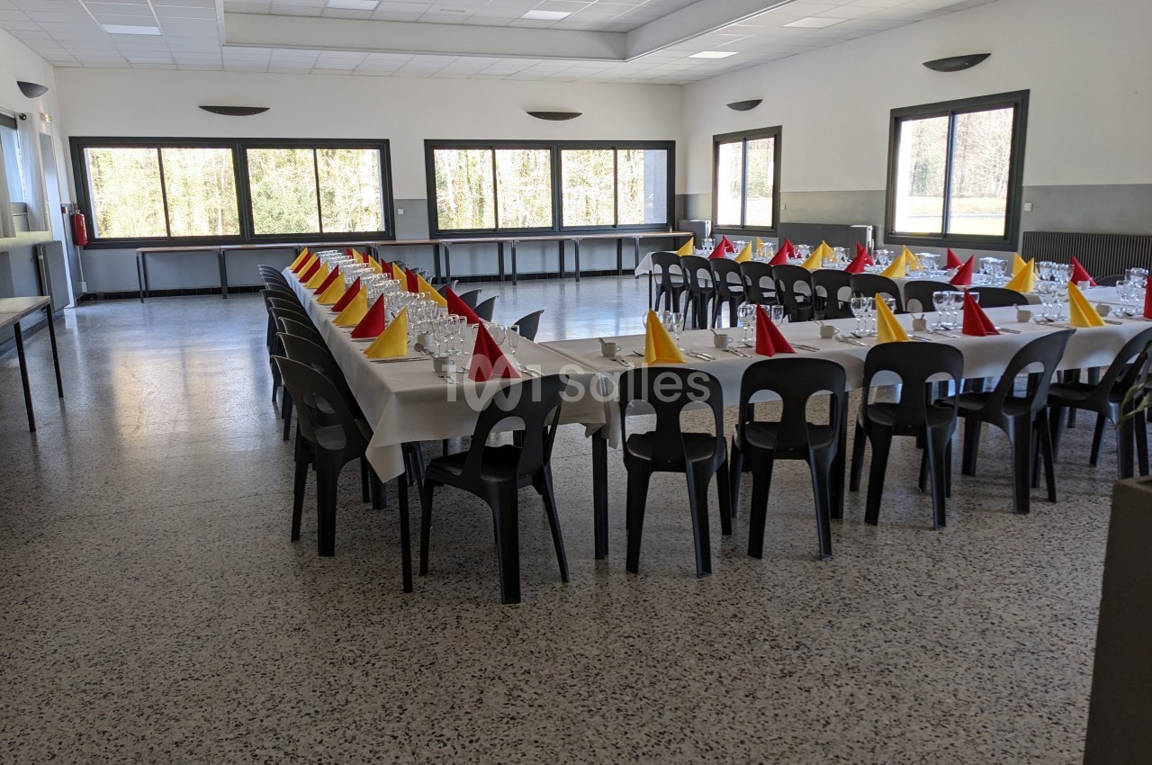 Salle de réception avec tables alignées, nappes blanches, chaises noires et serviettes pliées en rouge et jaune.