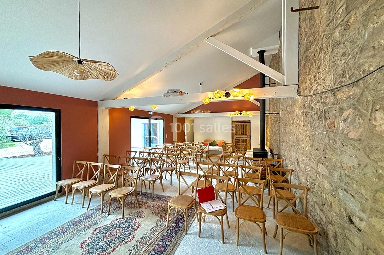 Salle lumineuse avec chaises en bois alignées, murs en pierre et fenêtres donnant sur une terrasse extérieure.