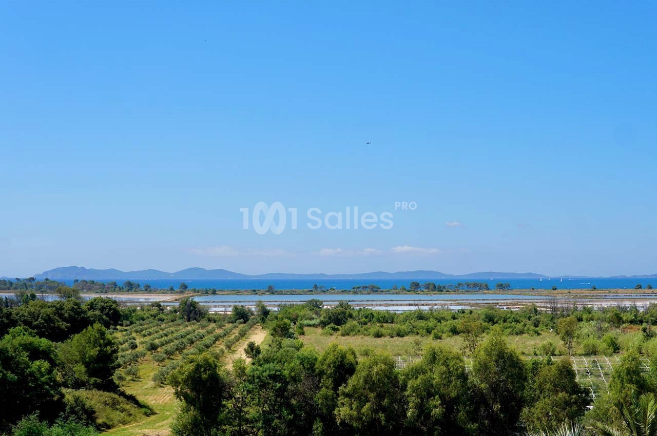 Paysage avec des champs verdoyants, des arbres, un lac en arrière-plan et des collines sous un ciel bleu dégagé.