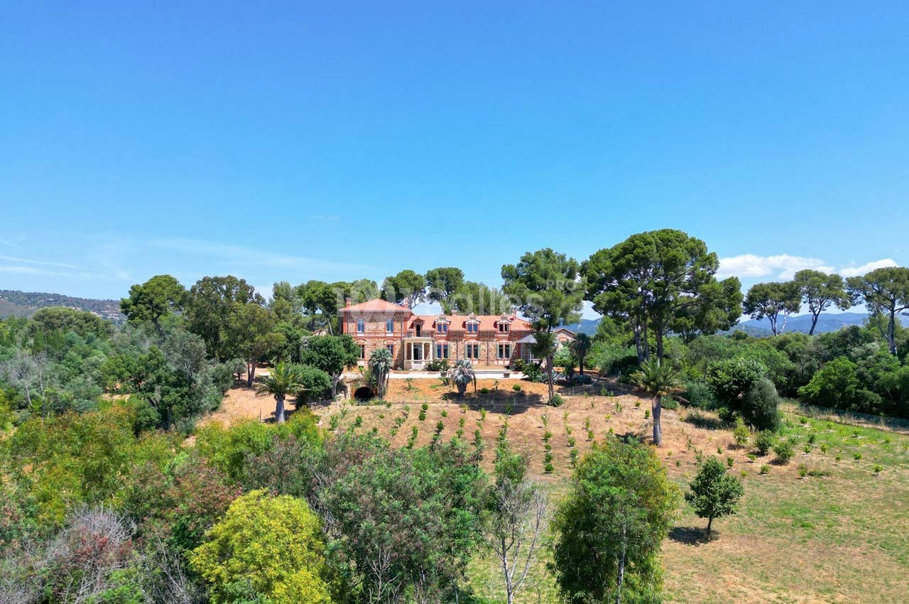 Vue aérienne d'une grande maison en pierre entourée de végétation et d'arbres, sous un ciel bleu clair.