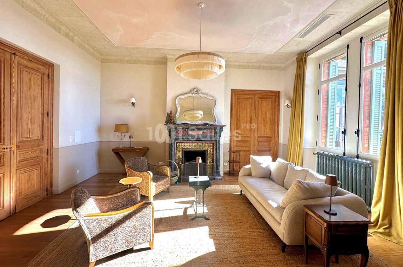 Salon lumineux avec cheminée décorative, mobilier élégant, grandes fenêtres et parquet en bois clair.
