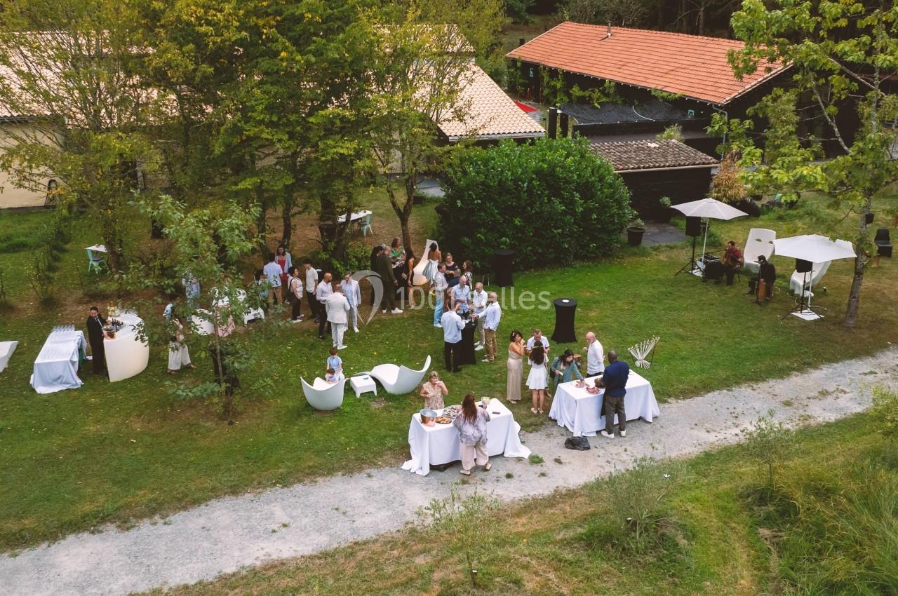 Vue aérienne d'un rassemblement en extérieur, avec des invités autour de tables dans un jardin verdoyant.