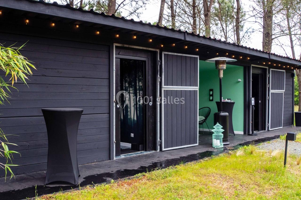 Façade d'un bâtiment moderne en bois noir avec des portes ouvertes, entouré de végétation et éclairé par des guirlandes.