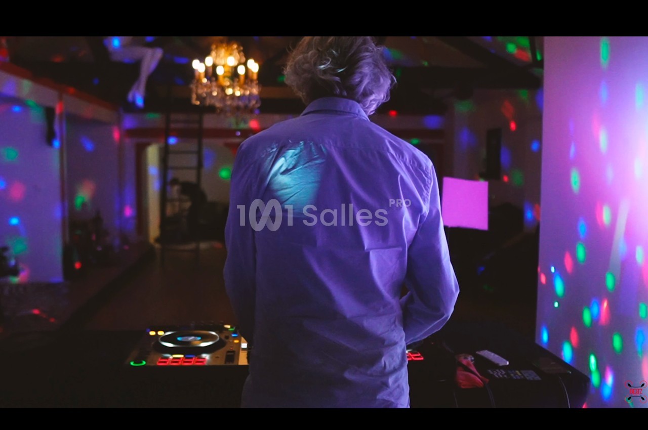 Un DJ vu de dos mixe dans une salle sombre éclairée par des lumières colorées et un lustre.