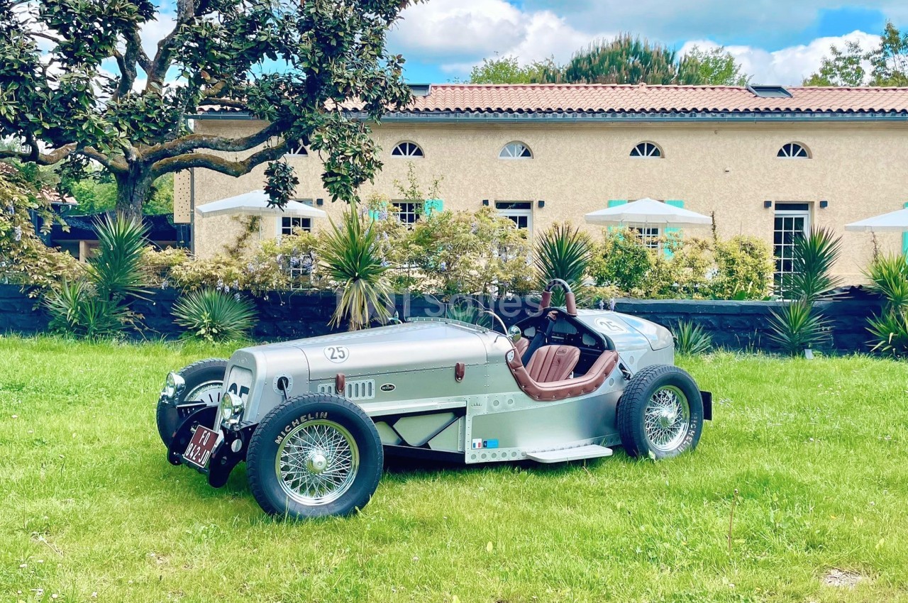Voiture de collection argentée stationnée sur une pelouse devant un bâtiment beige avec des parasols et des arbres.