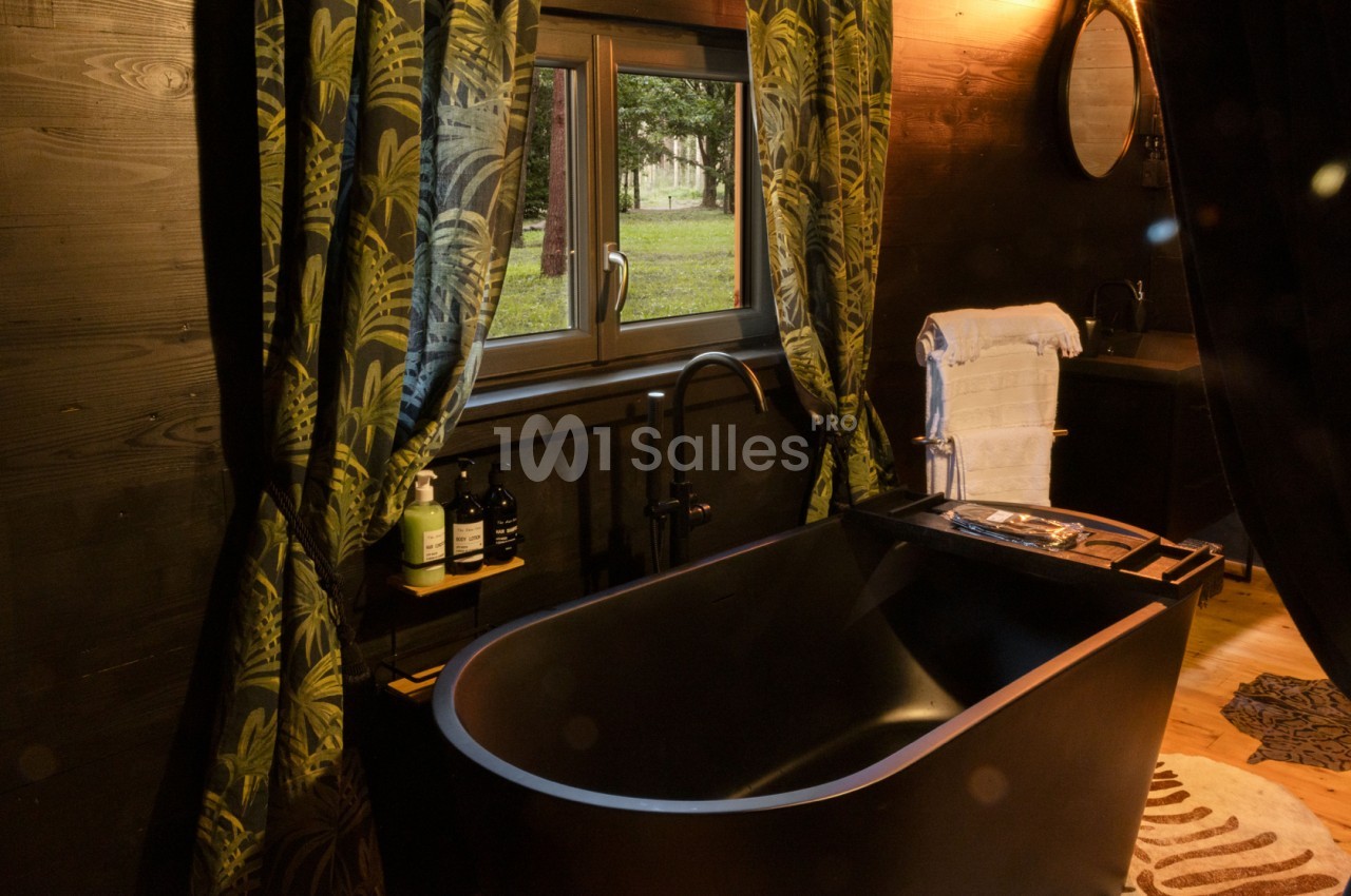 Baignoire noire dans une salle de bain en bois sombre avec rideaux à motifs végétaux et vue sur un jardin.