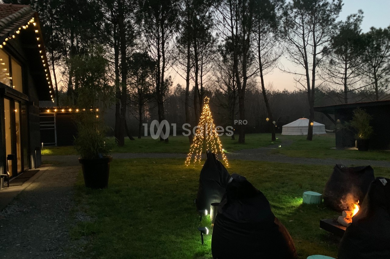 Cour extérieure au crépuscule avec un sapin lumineux, des poufs autour d'un brasero et des arbres en arrière-plan.