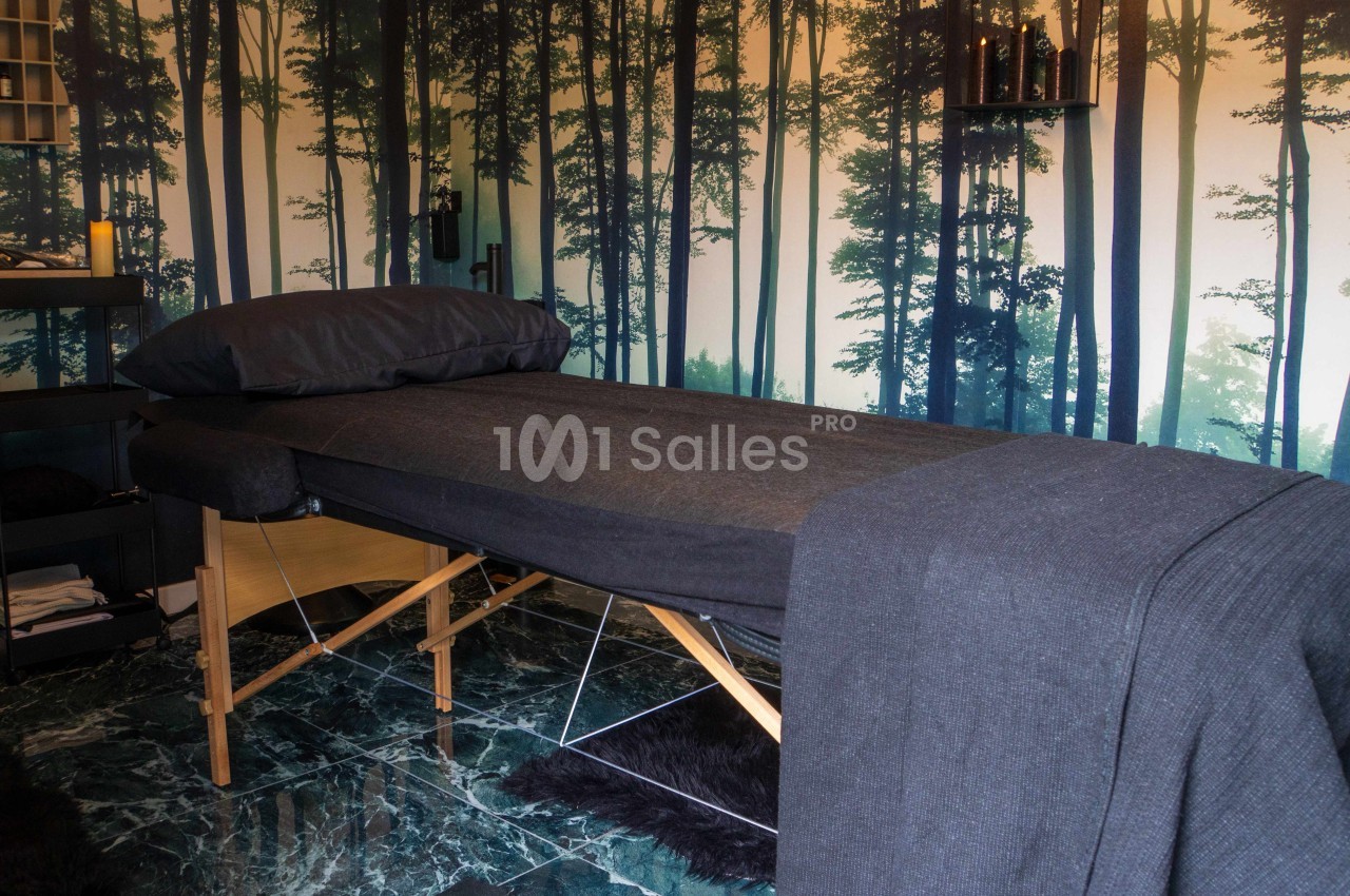 Table de massage recouverte de draps gris dans une pièce décorée d'un papier peint représentant une forêt.