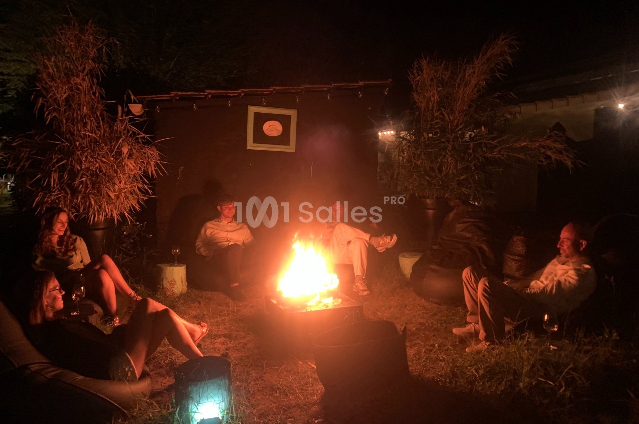 Personnes assises autour d'un feu de camp dans un jardin, éclairées par les flammes et des lumières tamisées.