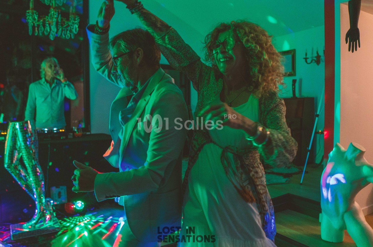 Un couple danse sous des lumières colorées dans une ambiance festive, avec un DJ en arrière-plan.