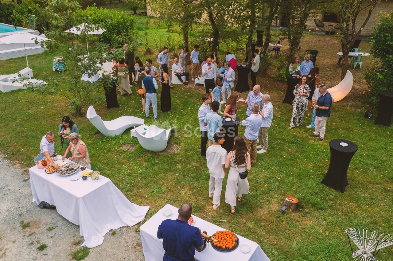 Groupe de personnes rassemblées dans un jardin pour un événement, avec tables de buffet et mobilier extérieur.