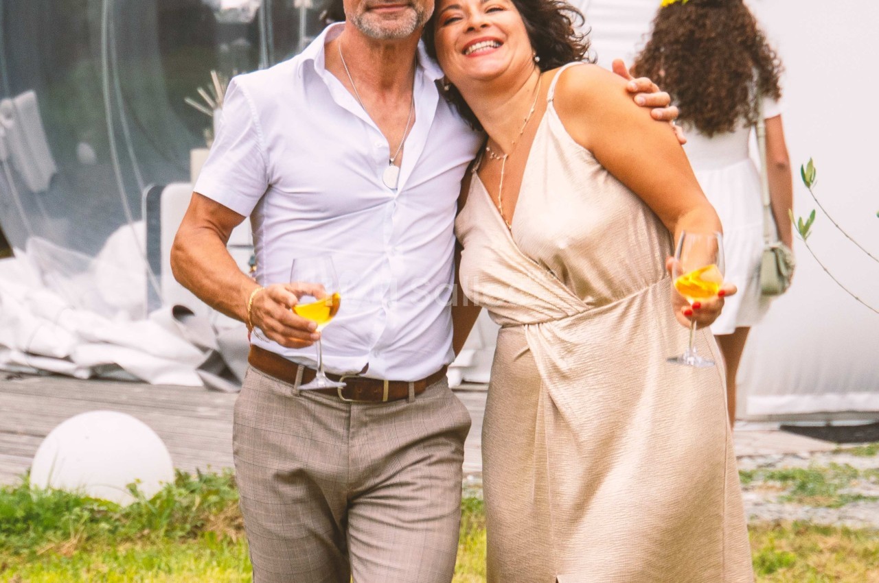 Un homme et une femme souriants tenant des verres, debout sur une pelouse devant une structure transparente.
