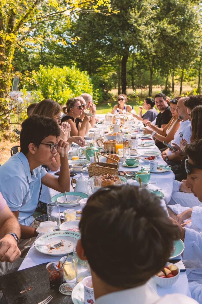 Groupe de personnes partageant un repas en plein air autour d'une longue table dans un cadre verdoyant.