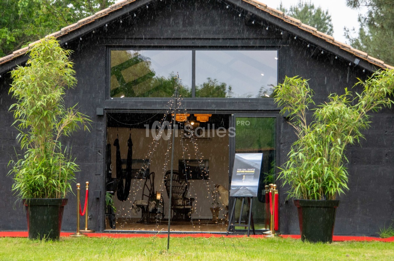 Façade d'un bâtiment noir avec de grandes baies vitrées, encadré par deux plantes en pot et éclairé à l'intérieur.
