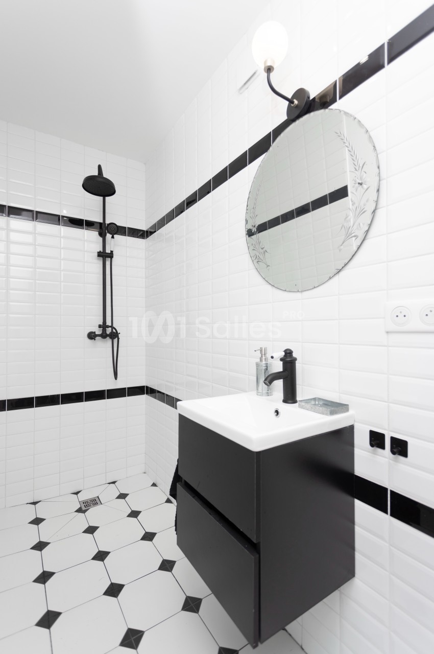 Salle de bain moderne avec carrelage blanc, douche noire, miroir rond et meuble vasque noir.