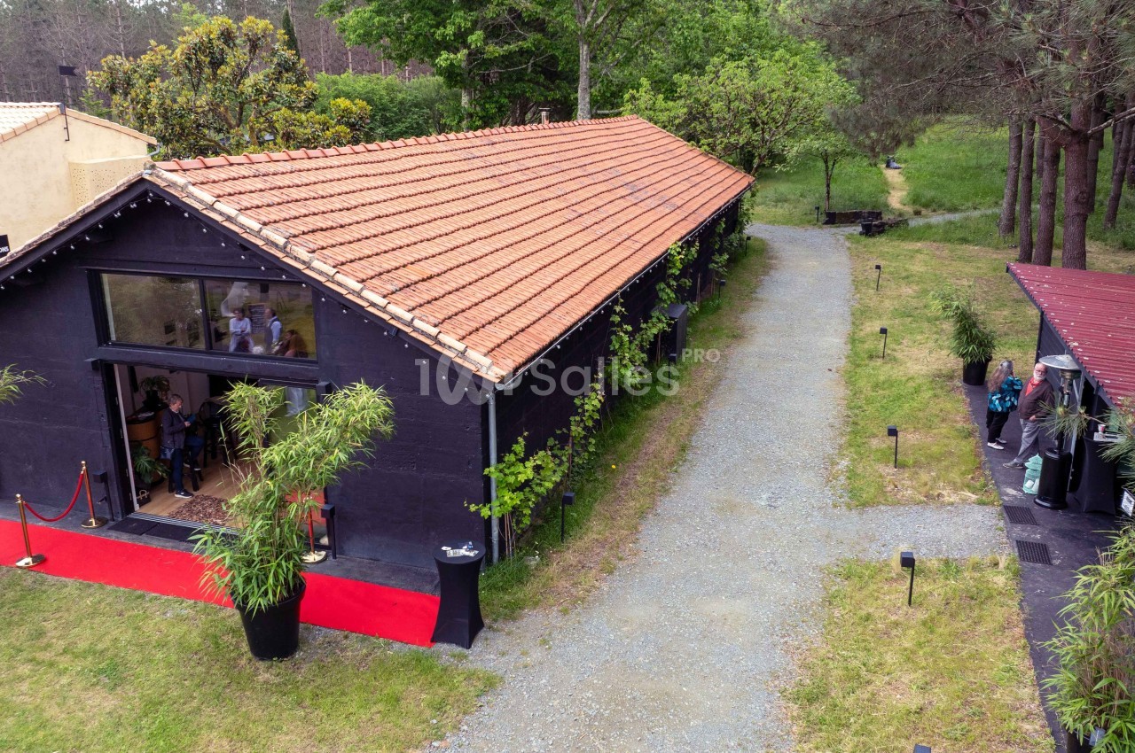 Bâtiment noir avec toit en tuiles rouges, entouré de végétation, avec une allée gravillonnée et une entrée vitrée.