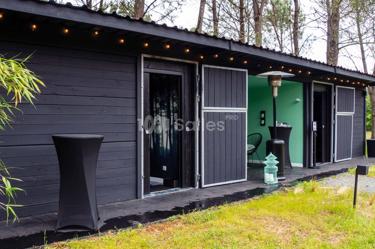 Façade d'un bâtiment moderne en bois noir avec des portes ouvertes, entouré de végétation et éclairé par des guirlandes.