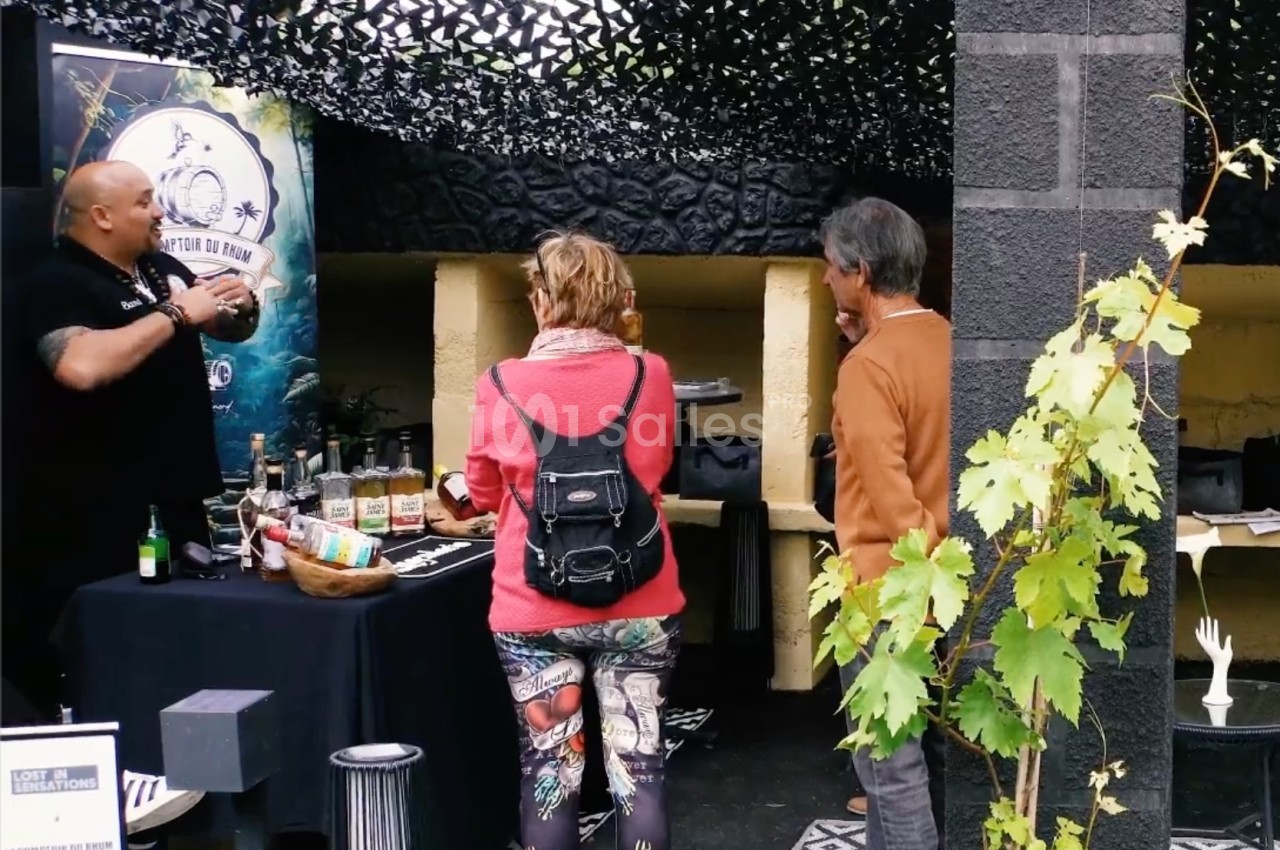Un stand de dégustation avec un exposant présentant des produits à deux visiteurs dans un espace extérieur abrité.
