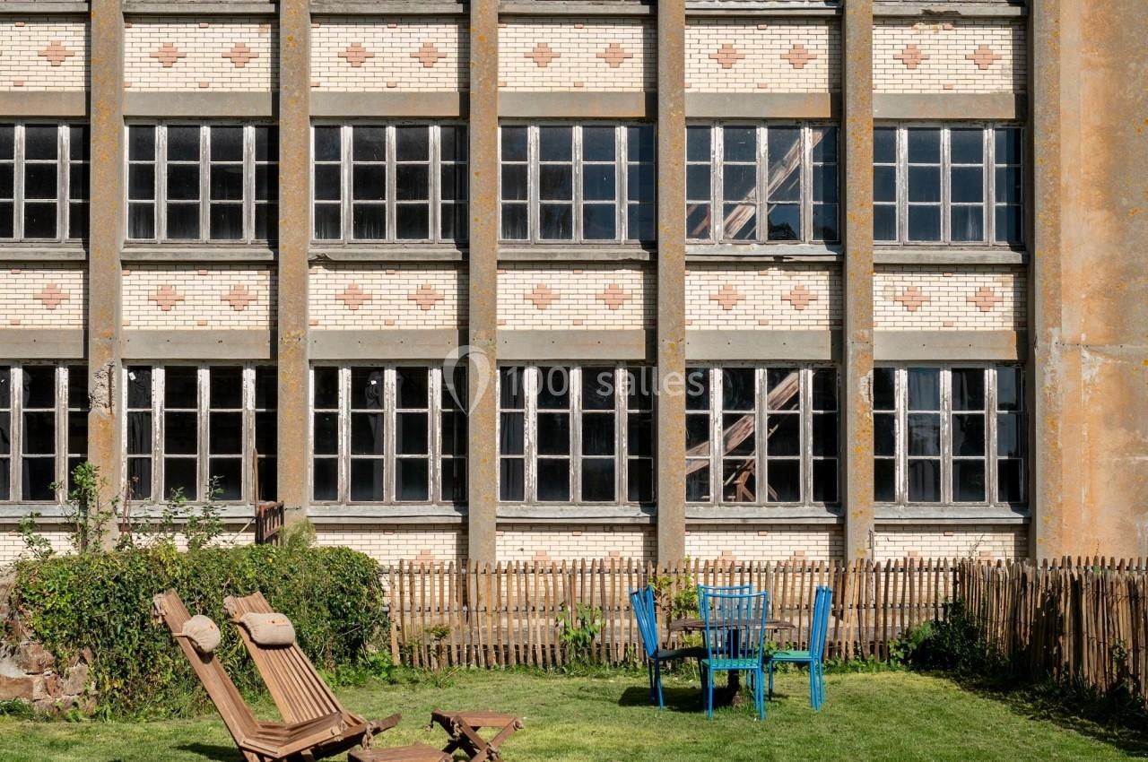 Façade d'un bâtiment ancien avec des fenêtres alignées, devant un jardin aménagé avec des chaises et des transats.