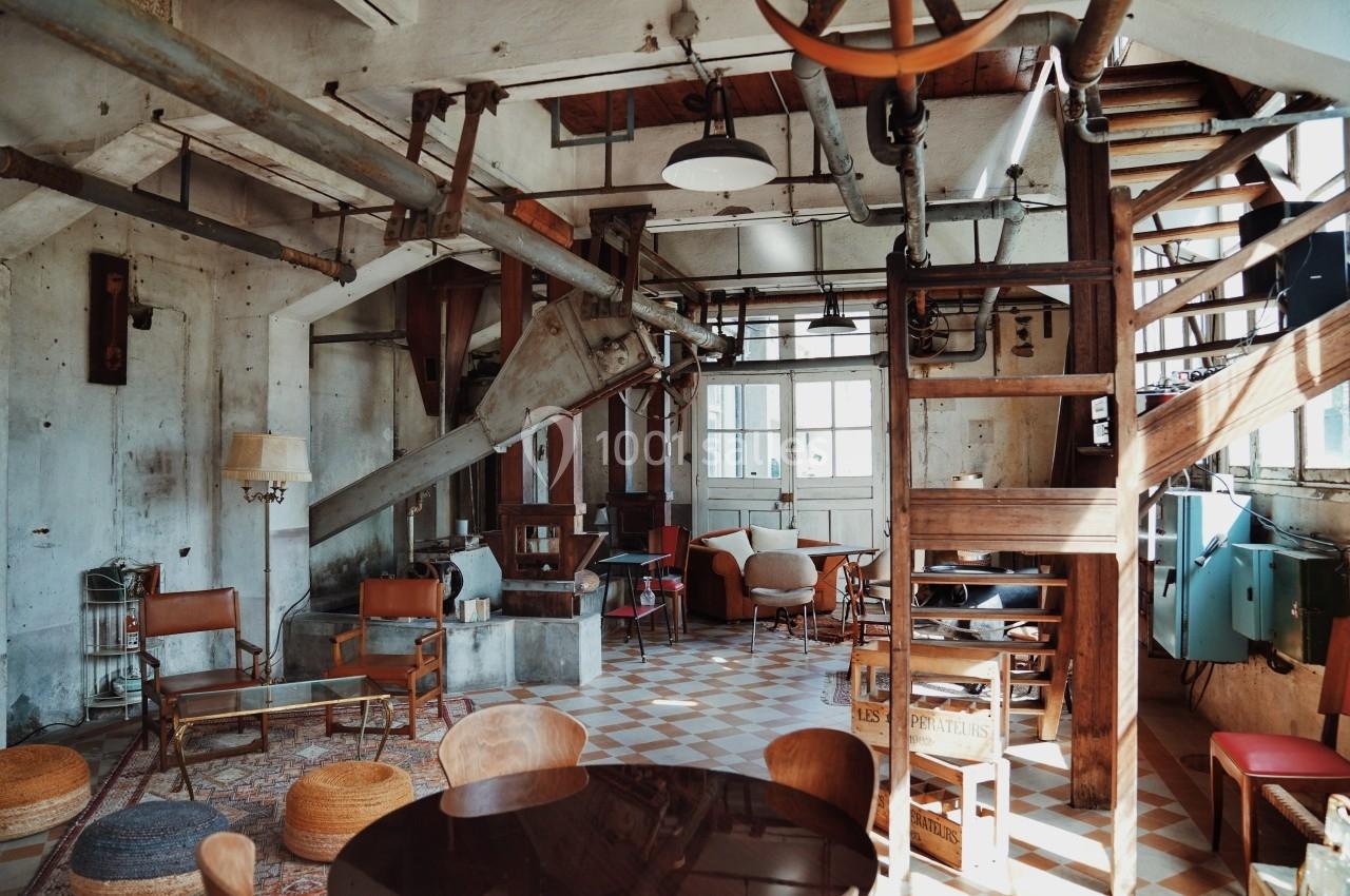 Intérieur d'un loft industriel avec meubles éclectiques, poutres apparentes et lumière naturelle entrant par des fenêtres.