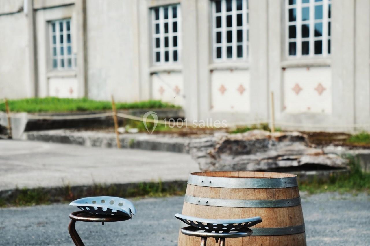 Deux tabourets en métal et un tonneau en bois utilisés comme mobilier extérieur devant un bâtiment industriel.