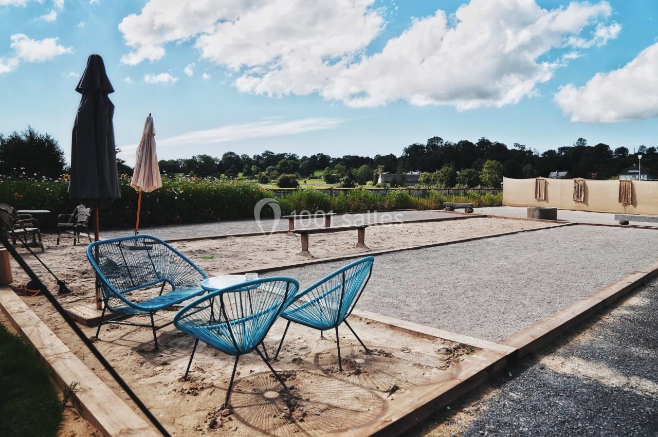 Terrain de pétanque en plein air avec chaises bleues et parasols, entouré de verdure sous un ciel partiellement nuageux.