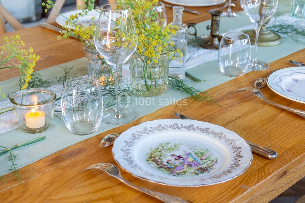 Table en bois dressée avec vaisselle vintage, verres, bougies et décoration florale champêtre sur un chemin de table vert.