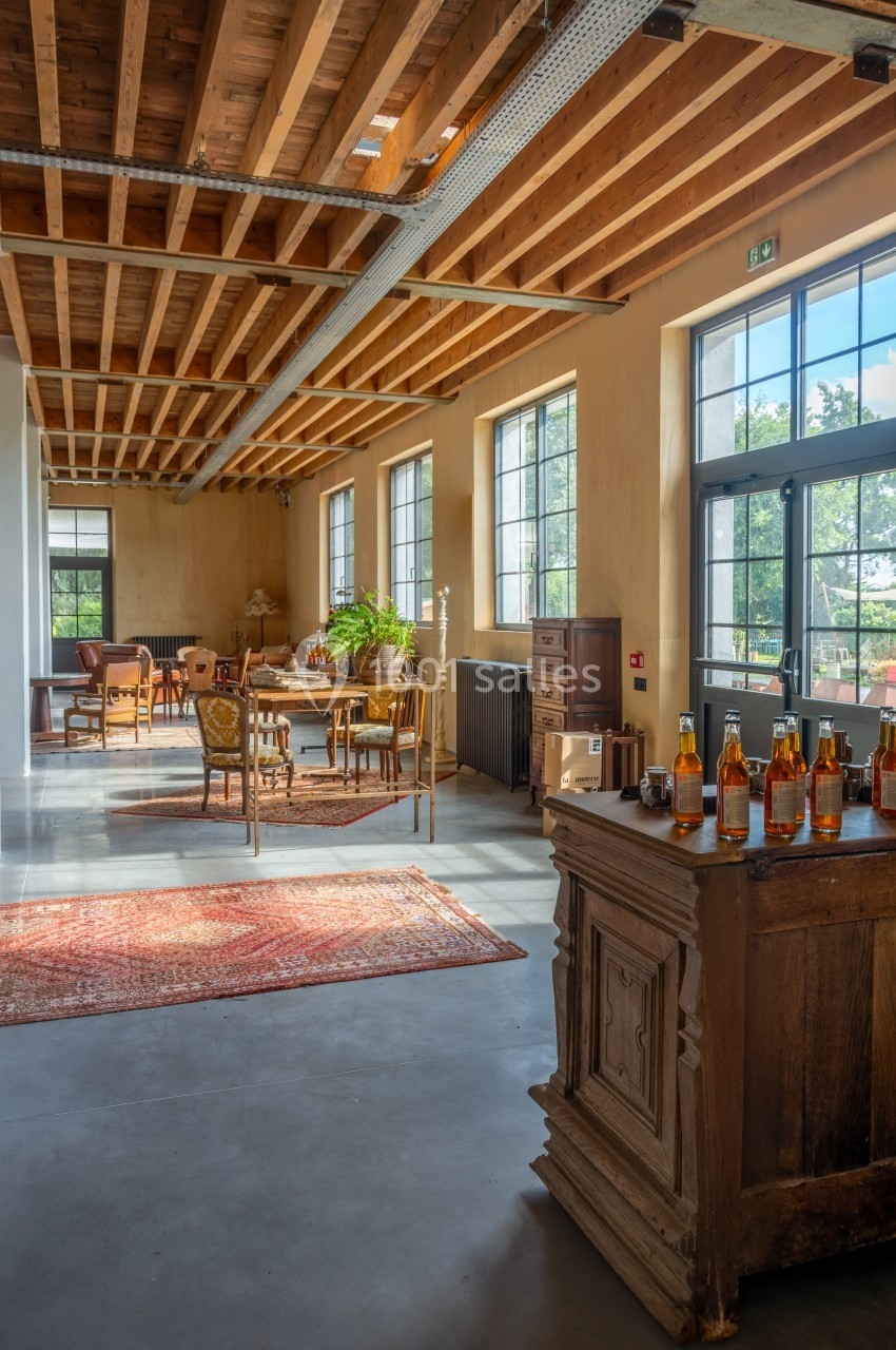 Intérieur lumineux avec plafond en bois, grandes fenêtres, mobilier ancien et bouteilles disposées sur un comptoir.