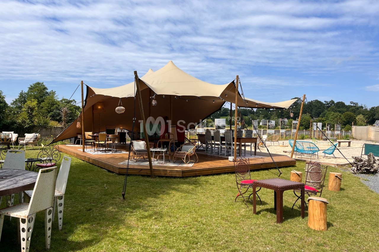Tente beige montée sur une terrasse en bois, entourée de tables et chaises dans un espace extérieur verdoyant.