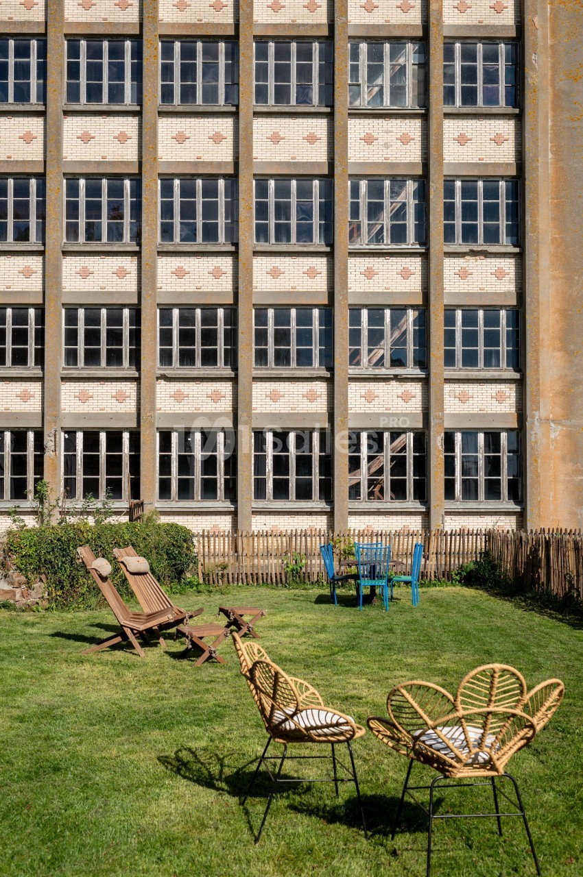 Façade d’un bâtiment en briques avec de grandes fenêtres, devant un jardin aménagé avec des chaises et des transats.
