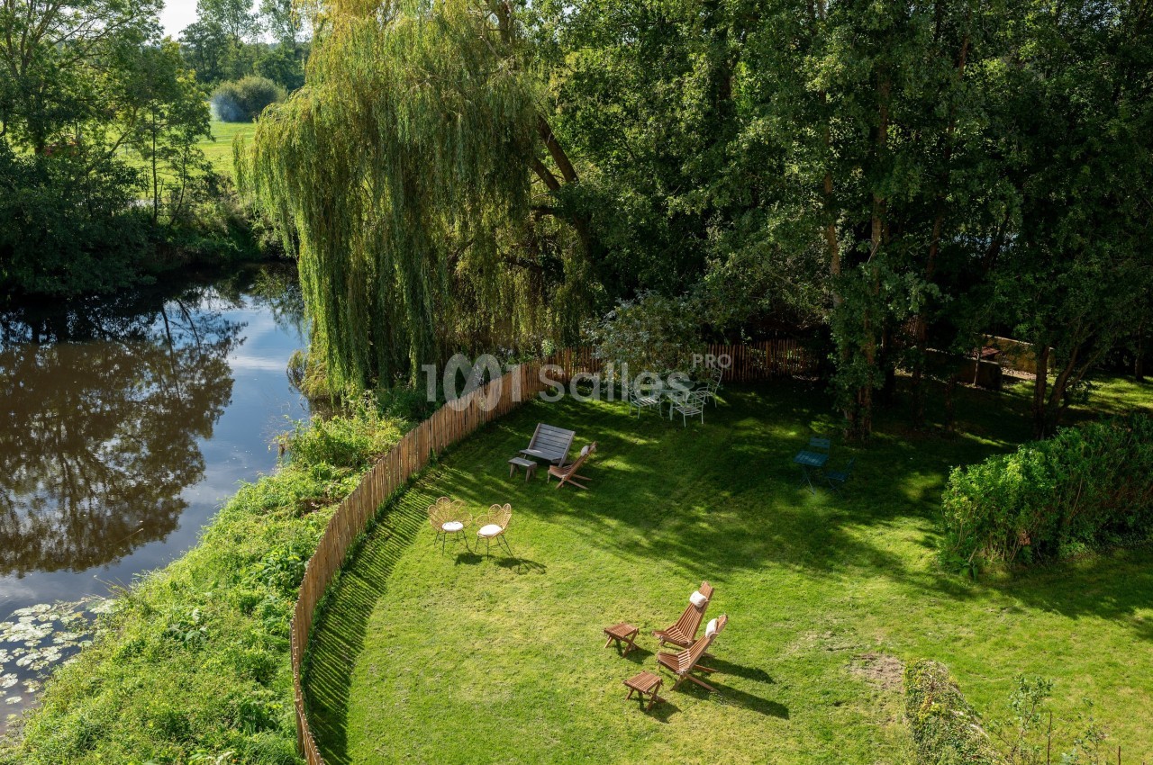 Jardin verdoyant avec chaises longues, table et rivière bordée d'arbres, entouré d'une clôture en bois.