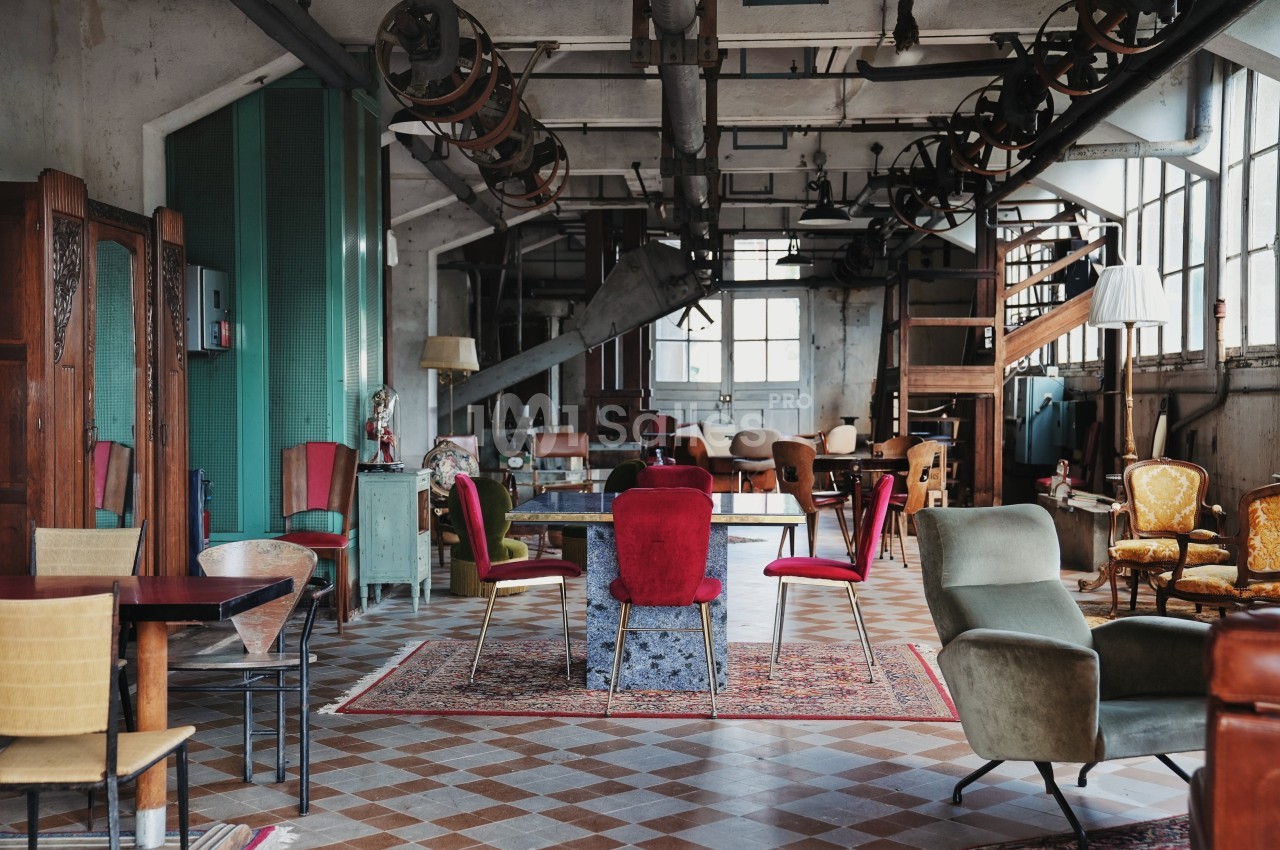 Salle industrielle aménagée avec des meubles éclectiques, incluant des chaises colorées et des tables sur un sol en carreaux.