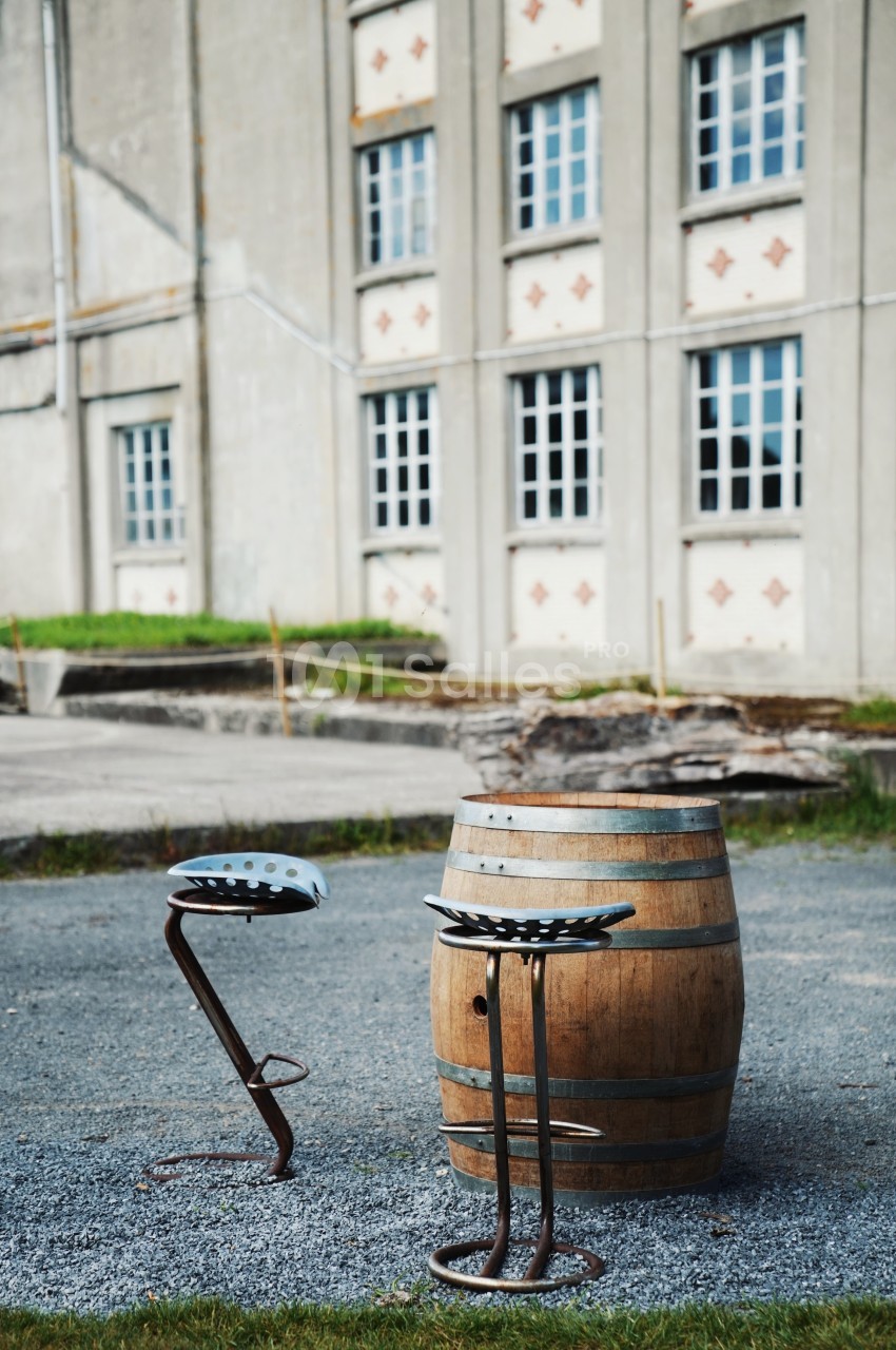 Deux tabourets en métal et un tonneau en bois disposés sur un sol gravillonné devant un bâtiment ancien.
