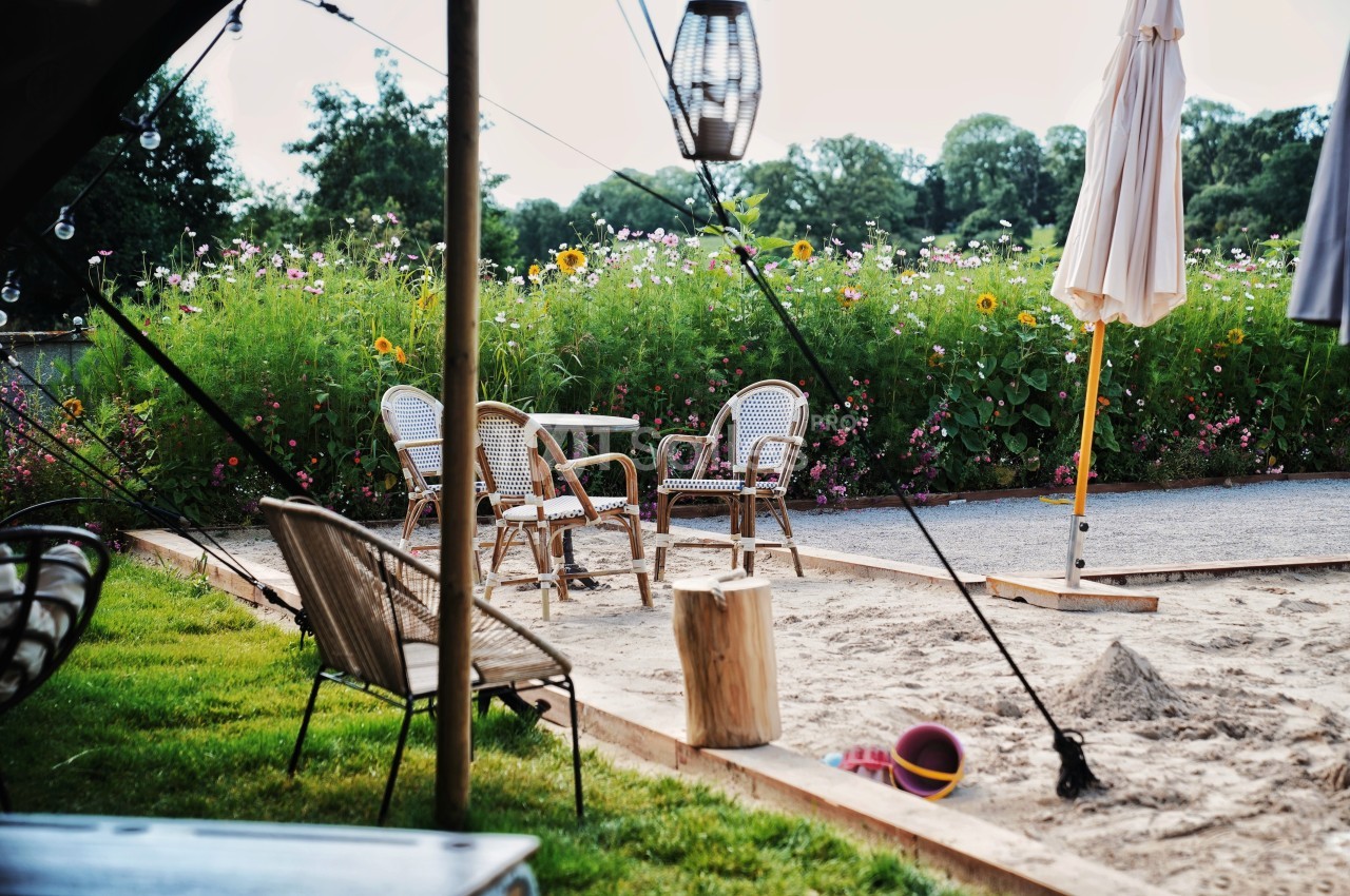 Espace extérieur avec chaises en rotin, table, parasol et jouets dans un bac à sable, entouré de fleurs et verdure.