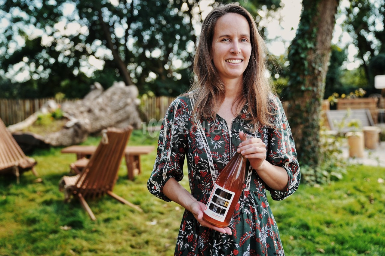 Une femme souriante tient une bouteille de boisson rosée dans un jardin avec des chaises en bois en arrière-plan.