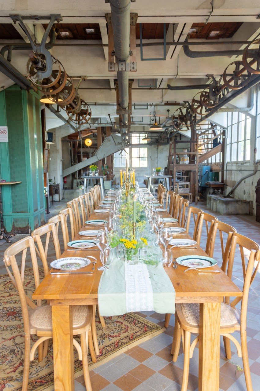 Table en bois longuement dressée avec vaisselle, bougies et fleurs, dans une salle industrielle lumineuse.