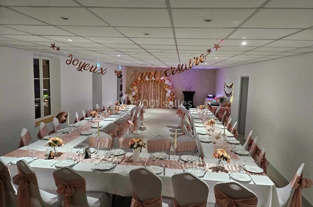Salle décorée pour un anniversaire avec tables dressées, chaises ornées de nœuds roses et guirlande ’Joyeux Anniversaire’.