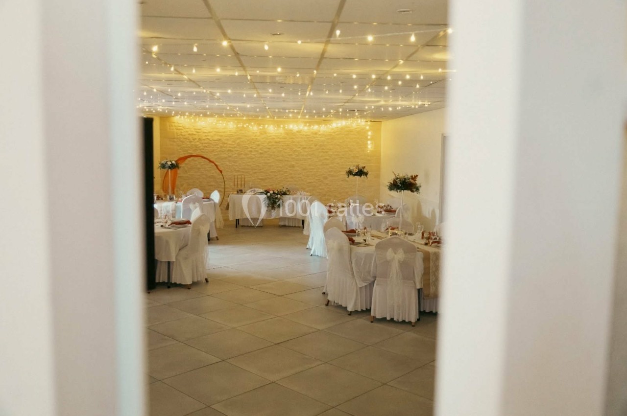 Salle de réception décorée avec des guirlandes lumineuses, des tables dressées et des chaises couvertes de housses blanches.