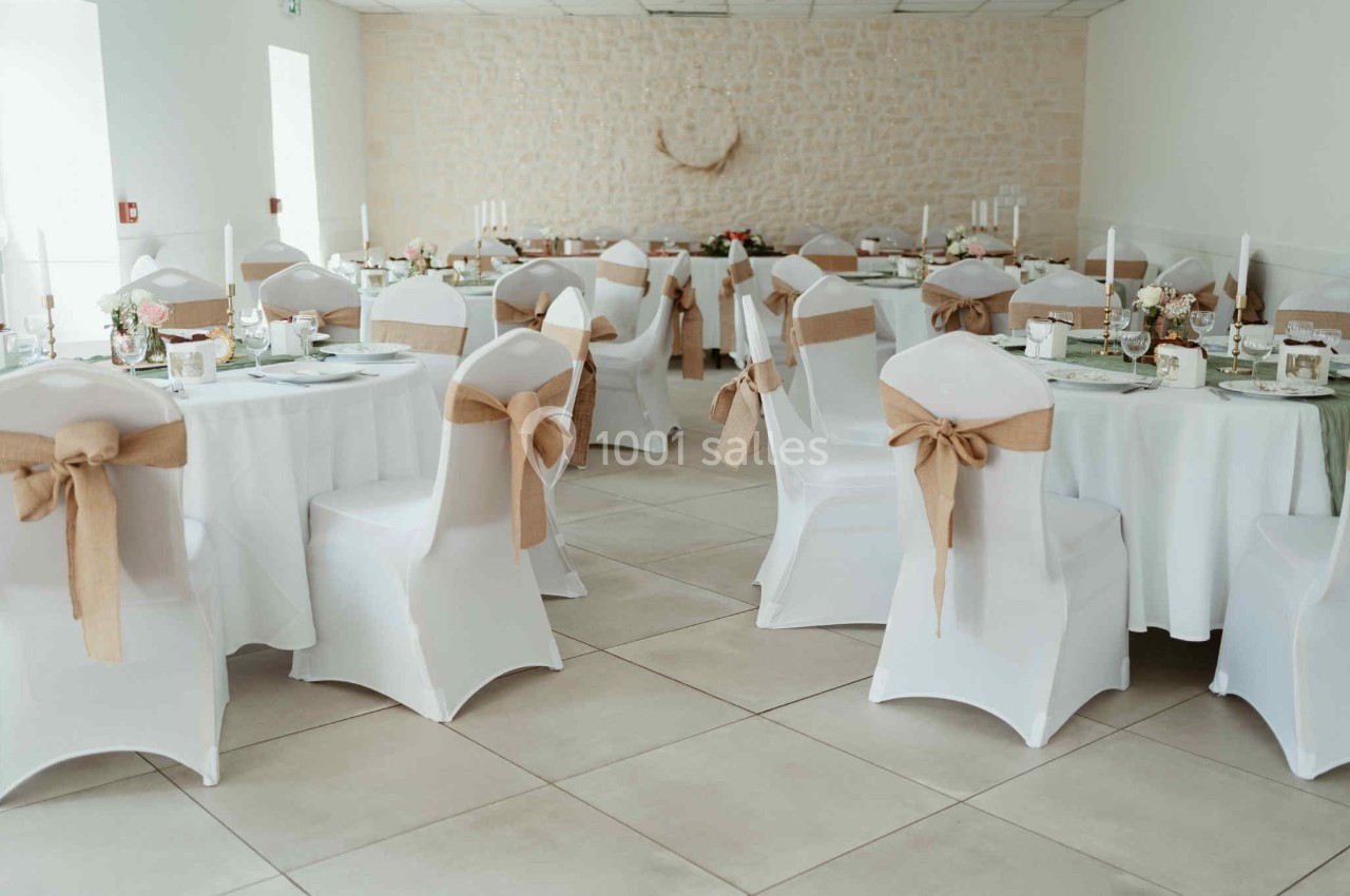 Salle de réception décorée avec des tables rondes, nappes blanches et chaises ornées de nœuds en toile de jute.