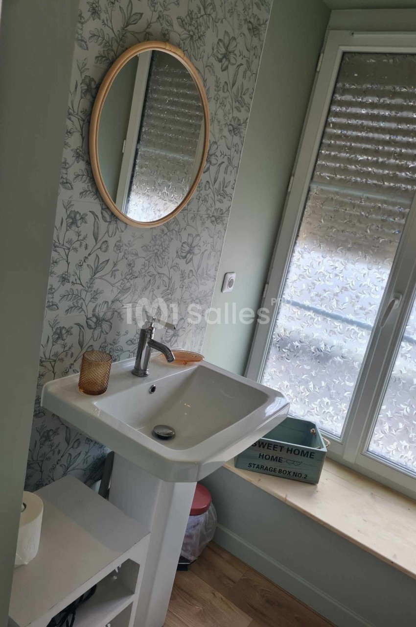 Lavabo blanc avec miroir rond, mur à motif floral, fenêtre avec stores baissés et étagère en bois.