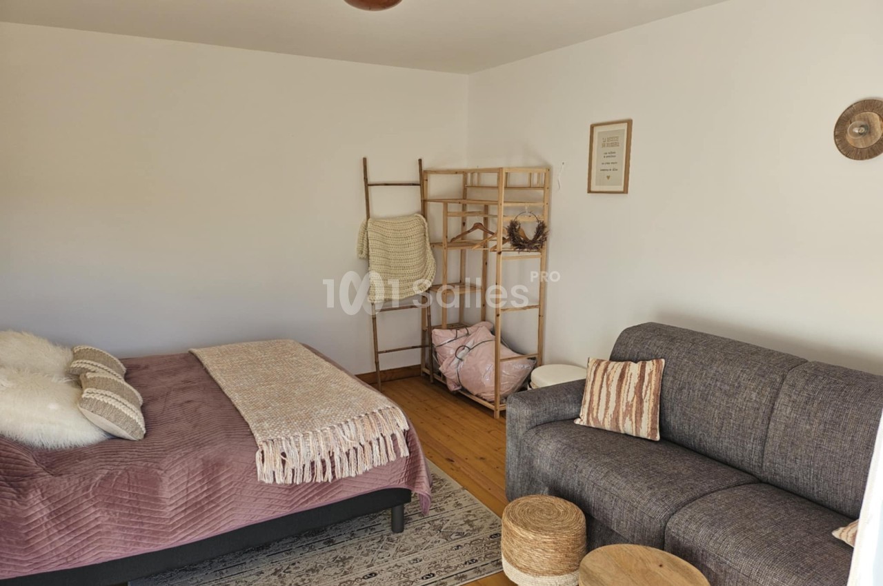 Chambre lumineuse avec lit double, canapé gris, étagère en bois, décor minimaliste et parquet au sol.