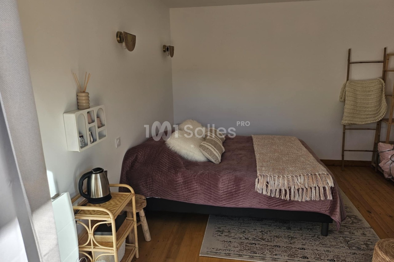 Chambre lumineuse avec lit double, plaid beige, coussins, petite étagère murale et coin avec bouilloire sur table en rotin.
