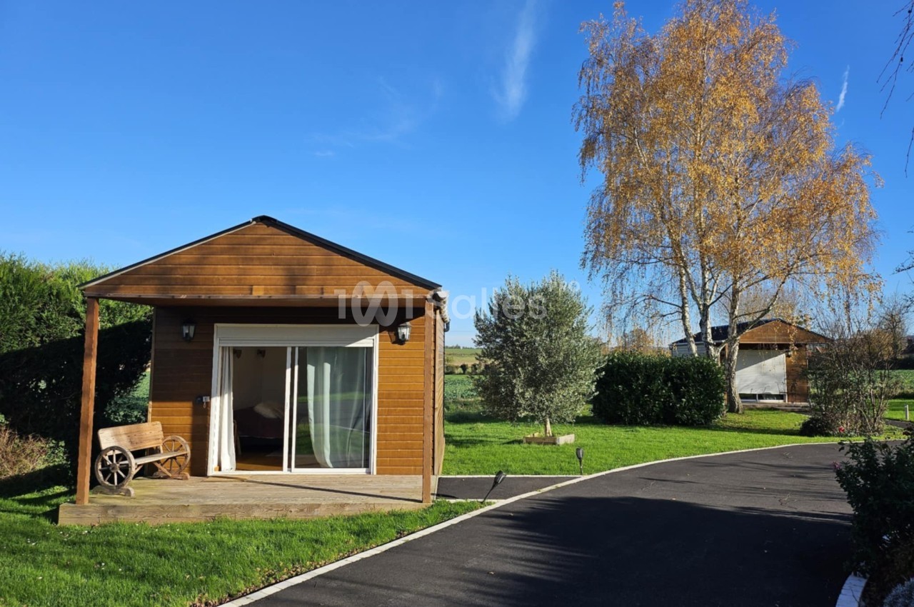 Chalet en bois avec terrasse, entouré de pelouse et d'arbres, sous un ciel bleu clair.