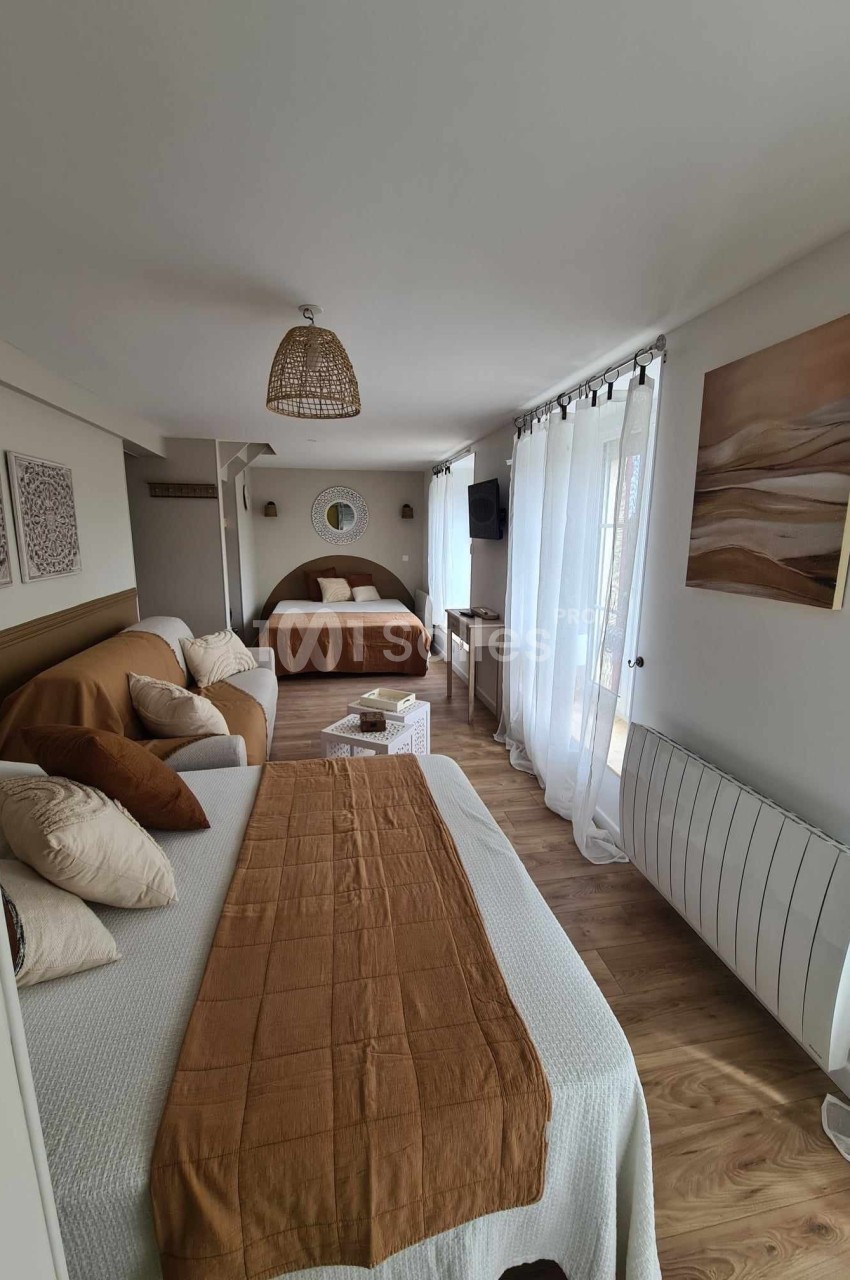 Chambre lumineuse avec deux lits, canapé, décorations murales, parquet et grande fenêtre avec rideaux blancs.