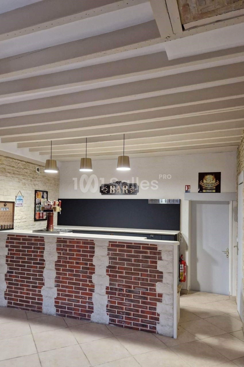 Comptoir de bar en briques avec panneau ’BAR’, trois suspensions lumineuses et murs en pierre apparente.