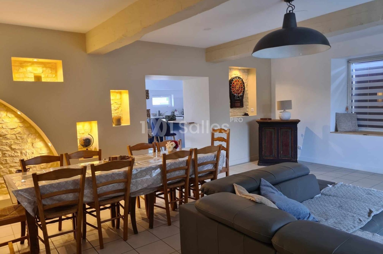 Salle à manger avec table en bois, chaises, murs en pierre éclairés, canapé en cuir et vue sur une cuisine ouverte.