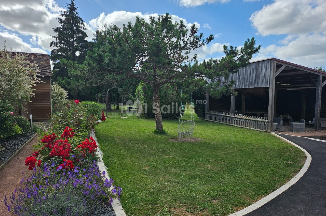 Jardin avec pelouse, fleurs colorées, arbre central, fauteuil suspendu et abri en bois sous un ciel partiellement nuageux.